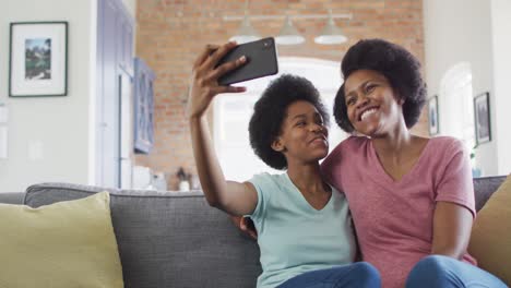 Happy-african-american-mother-and-daughter-sitting-on-sofa-using-smartphone,-taking-selfie