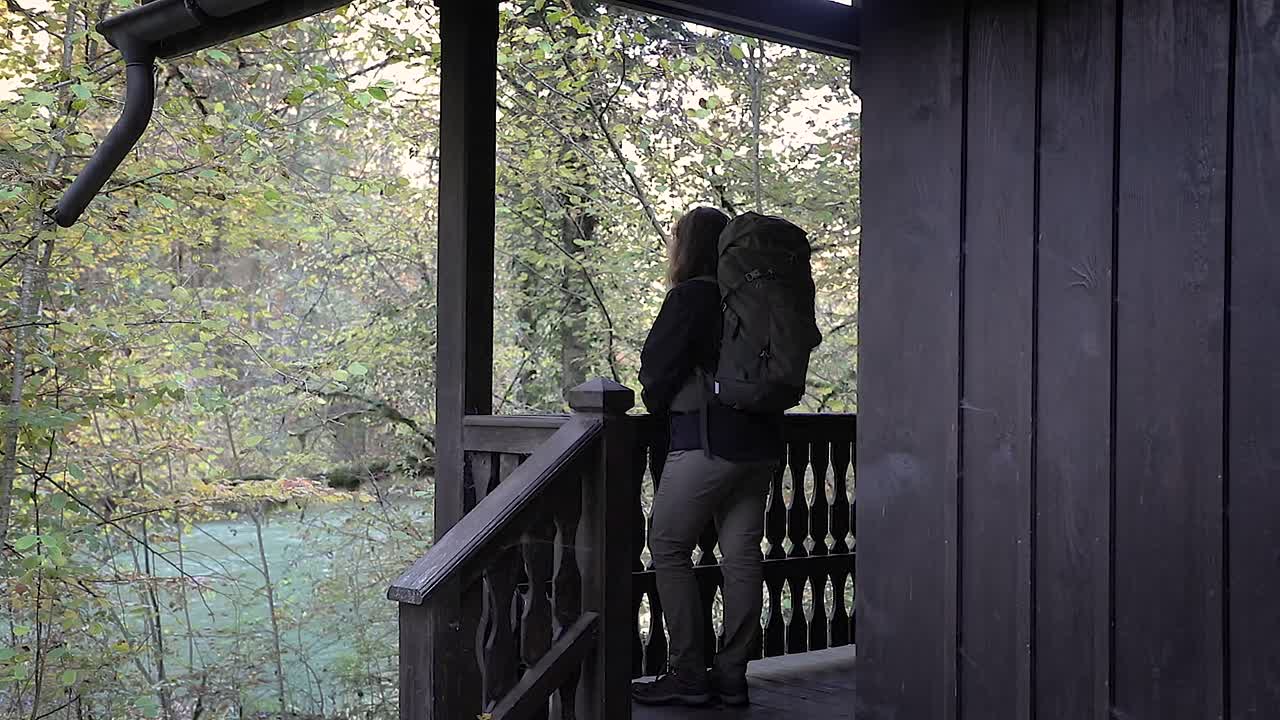 Girl backpacker standing alone on wooden cabin porch, looking out at scenery