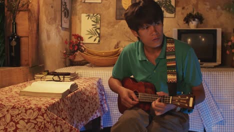 a young asian male playing a ukulele while sitting on a chair in a rustic room