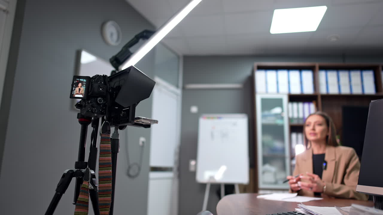 Woman in beige jacket lit by the day-light lamp is filmed by a professional camera. Female speaker records blog content. Low angle view.
