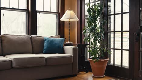 a-couch-in-a-sun-room-with-a-blue-pillow-on-it-sitting-next-to-a-large-plant,-lamp,-and-large-windows-letting-in-natural-sunlight