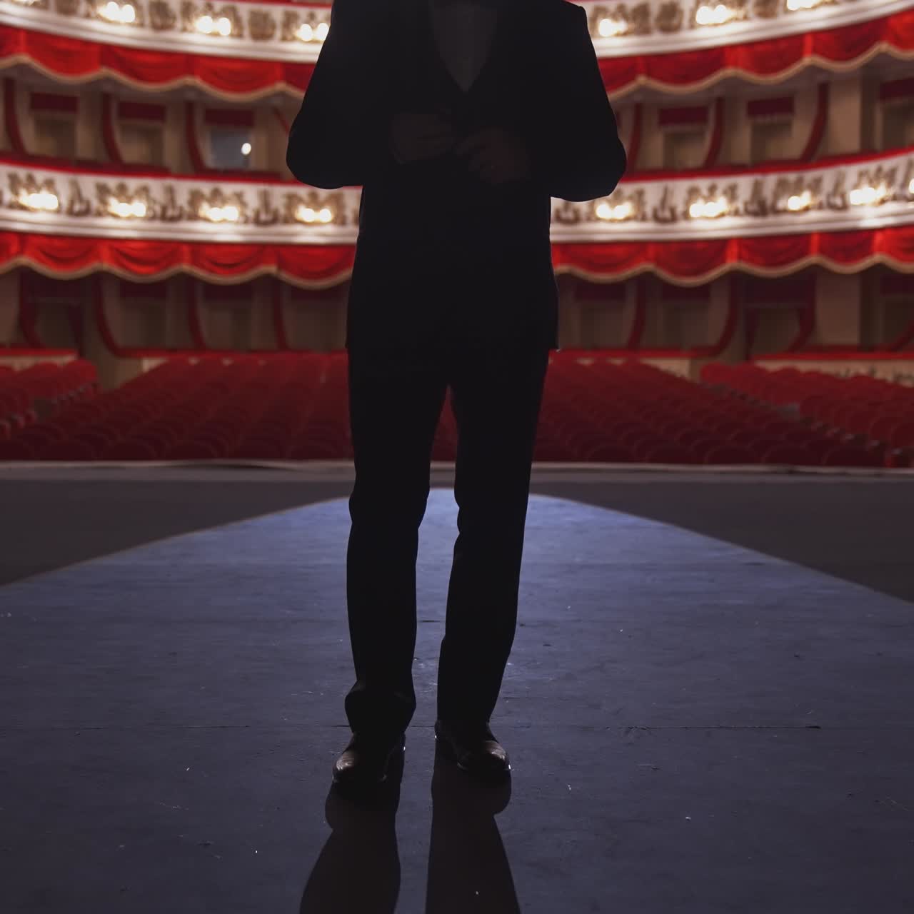 Man in suit standing on stage back to auditorium. Dark silhouette of unrecognizable actor on the background of classic theatre hall in red and white colors.