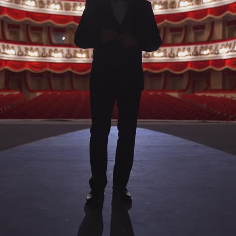 man in suit standing on stage back to auditorium. dark silhouette of unrecognizable actor on the background of classic theatre hall in red and white colors.
