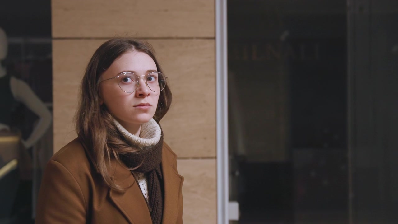 Una signora si ferma davanti a un negozio con vetri riflettenti. Scena serale con una donna in cappotto marrone e bagliore al neon. Scena di centro commerciale illuminato a giorno con una donna che esita fuori da una vetrina con manichini.