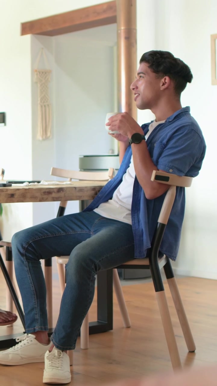 Video vertical: Hombre con camisa azul apoyado, usando una tablet, girando, respondiendo y bebiendo de una taza en casa.
