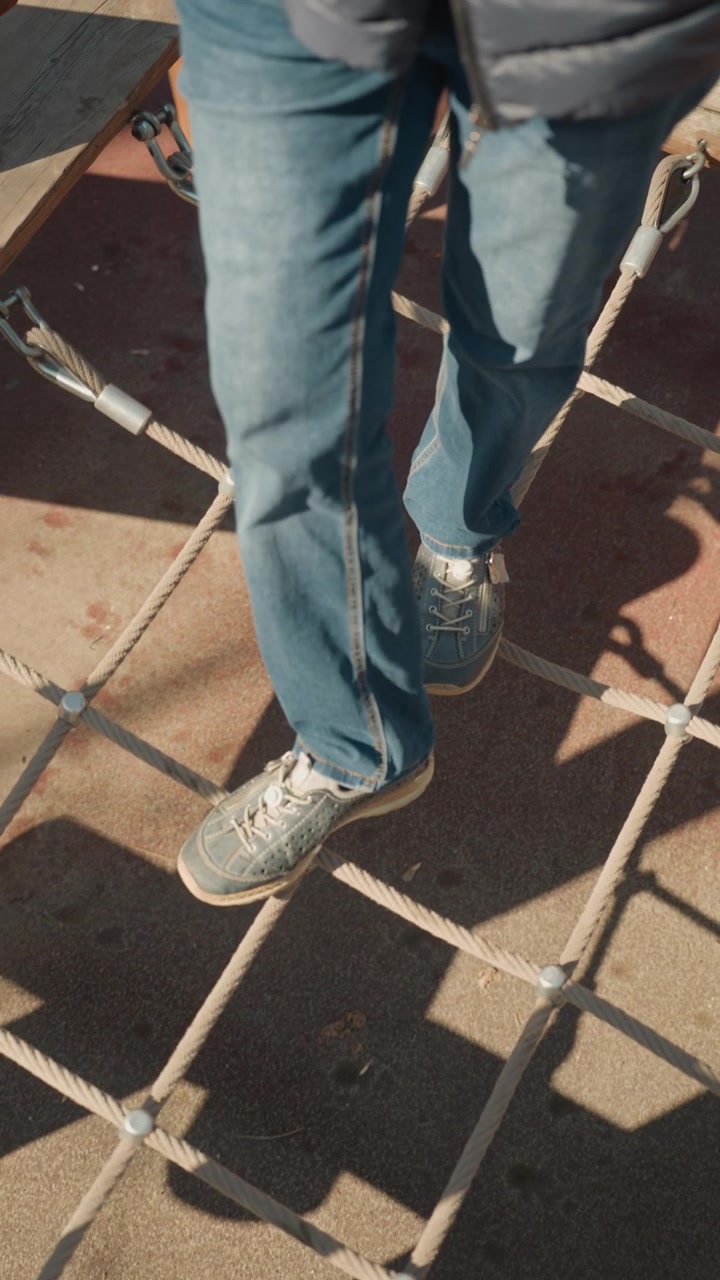 Close up of person legs and feet walking across outdoor rope bridge with wooden planks. person wears blue jeans gray sneakers and scene features sunlight casting shadows on concrete ground