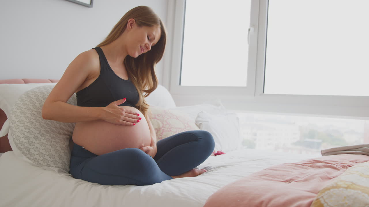 Pregnant Woman Wearing Fitness Clothing Sitting On Bed At Home Holding Stomach Feeling Baby Kick