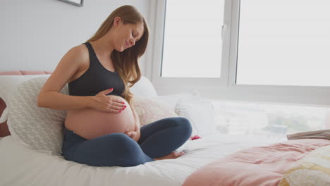 pregnant woman wearing fitness clothing sitting on bed at home holding stomach feeling baby kick