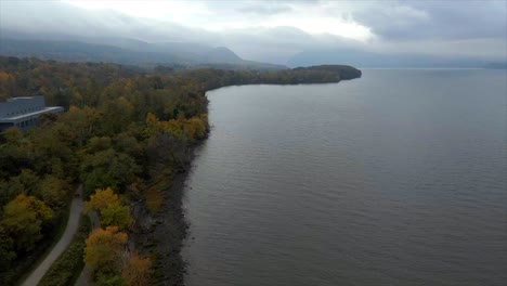 Fliegen-Entlang-Der-Küste-In-Der-Nähe-Eines-Farbenfrohen-Herbstwaldes-Mit-Dramatischen-Wolken-Und-Bergen-In-Der-Ferne