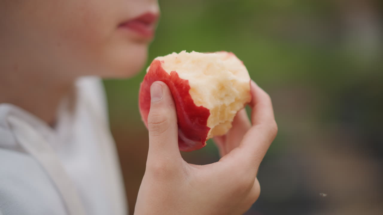 Closeup white girl biting apple outdoors, tight crop on mouth and hand, white hoodie, juicy bite with red peel contrast, soft bokeh background, mindful eating moment, afterschool snack vibe, cinematic