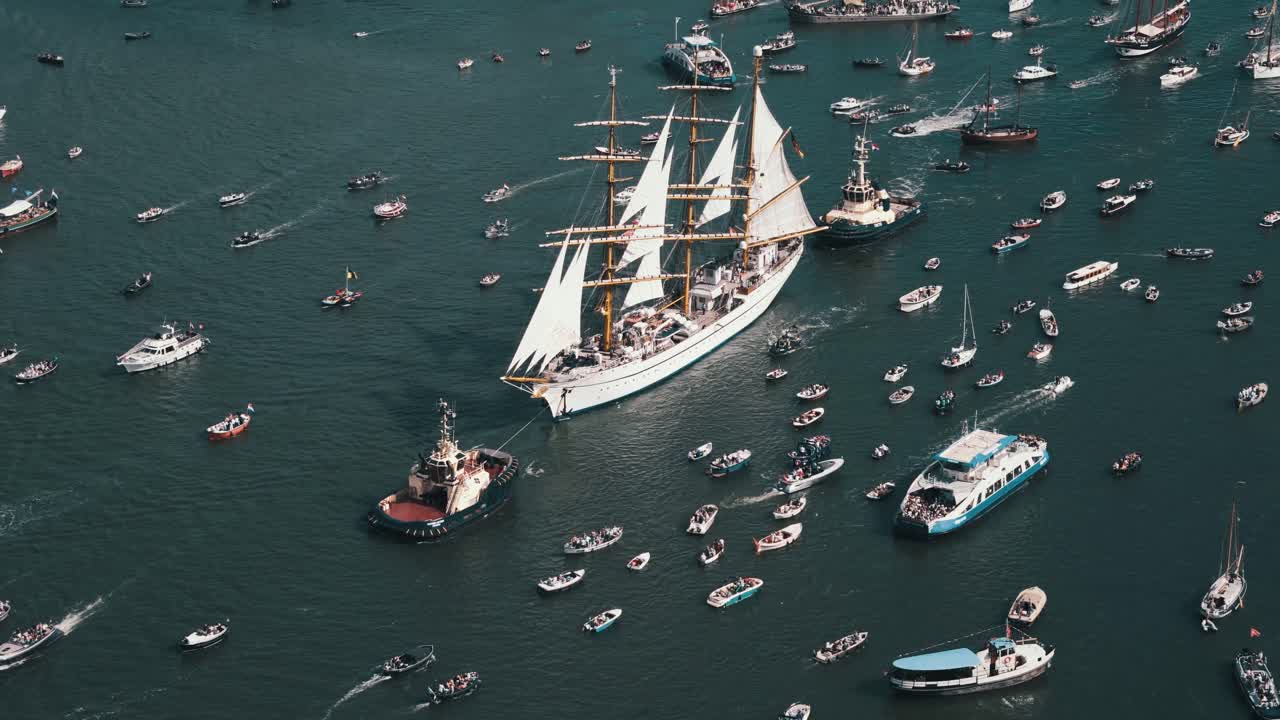 Drone overview of tug boat towering large white sailboat during busy Amsterdam Sail Festival in clear daylight