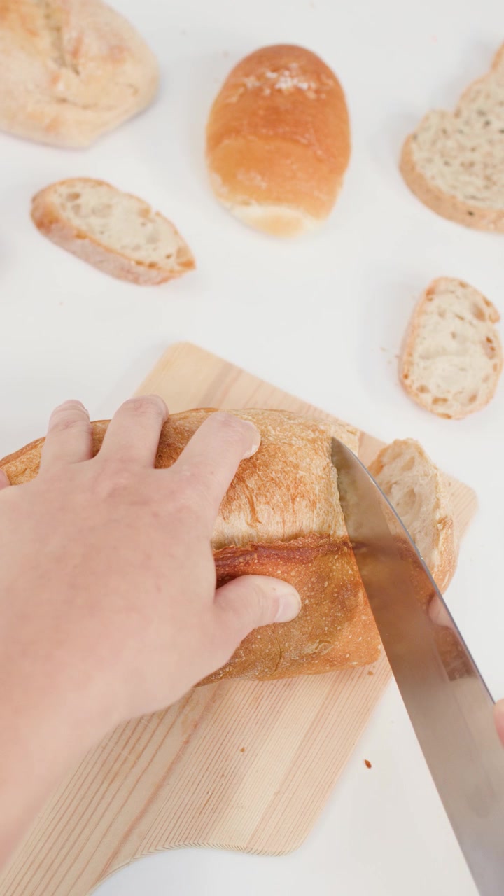 Top view of person cutting artisan bread with knife on wooden board against white surface.