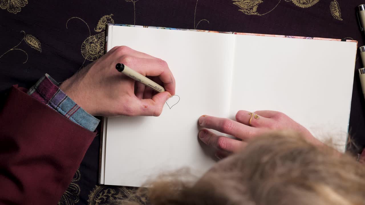 Young fine art illustrator graphic designer artist drawing a heart with a fineliner in his sketchbook - close up top shot - 4k