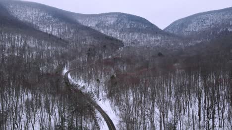 Flying-over-a-snowy-mountain-valley-with-forests-below-and-a-remote-mountain-highway,-and-snow-covered-mountains-in-the-distance