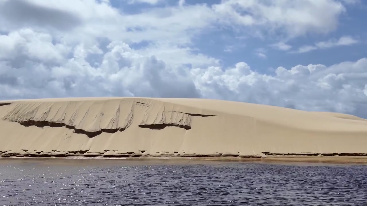 Aufnahme aus der Froschperspektive von einem Boot auf dem Fluss Preguiça, mit großen Küstendünen und Windturbinen unter bewölktem Himmel in Maranhão, Brasilien.