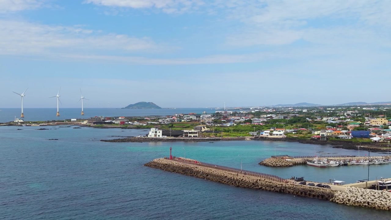済州島新昌里港上空からの空撮プルバック。防波堤、漁船、洋上風力タービン、そして沿岸の村が姿を現す。