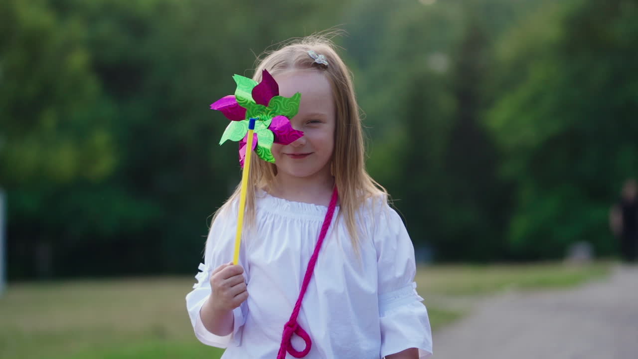 Lovely girl posing to camera. Positive little girl closing her face with toy windmill in the park. Nice child with colorful toy outdoors in summer.