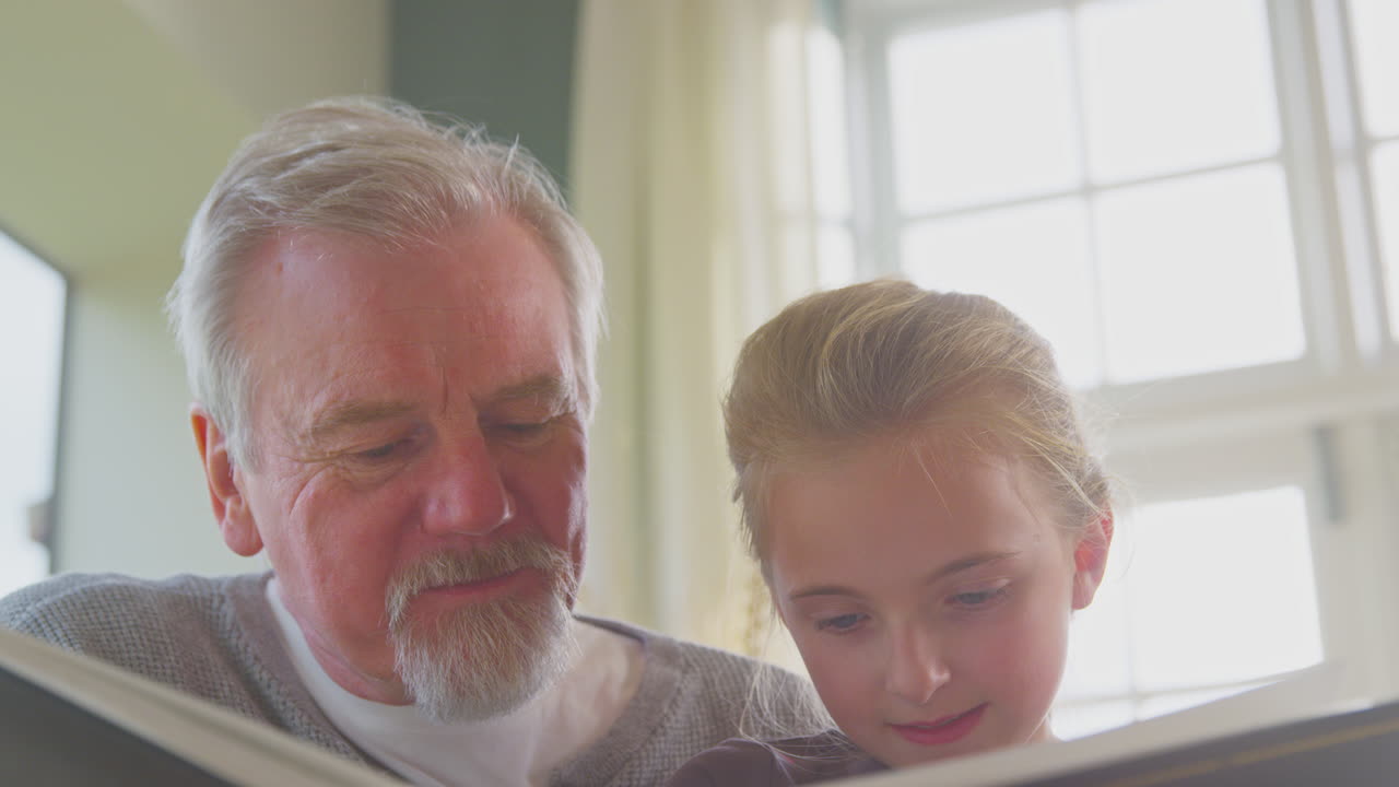 Close Up Of Grandfather And Granddaughter Looking Through Photo Album In Lounge At Home Together