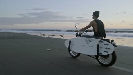 mężczyzna w wełnianym kapeluszu i okularach przeciwsłonecznych jadący motocyklem po plaży z deską surfingową u boku