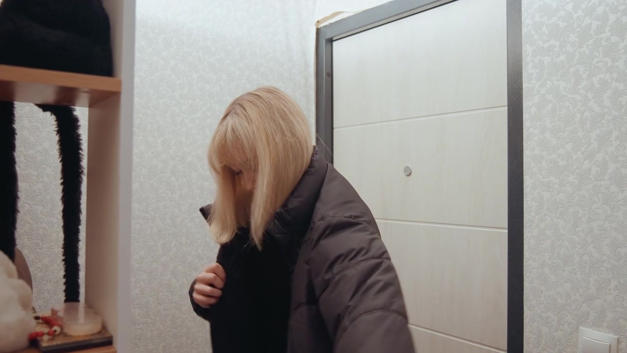 Young white woman putting on coat near apartment door, slipping into puffer jacket, checking pockets, zipping up, gazing down, commuter returning from work, student grabbing keys, subdued filmic