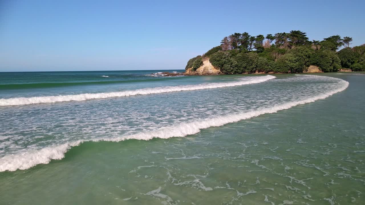 Um drone aéreo voa perto da costa da Rawera Beach em um dia ensolarado, capturando ondas, a costa arenosa e um promontório coberto de árvores no plano de fundo da Nova Zelândia