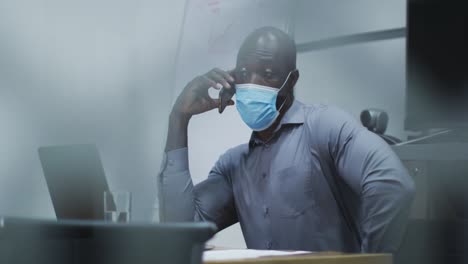 african american businessman in face mask talking on smartphone, using laptop in conference room