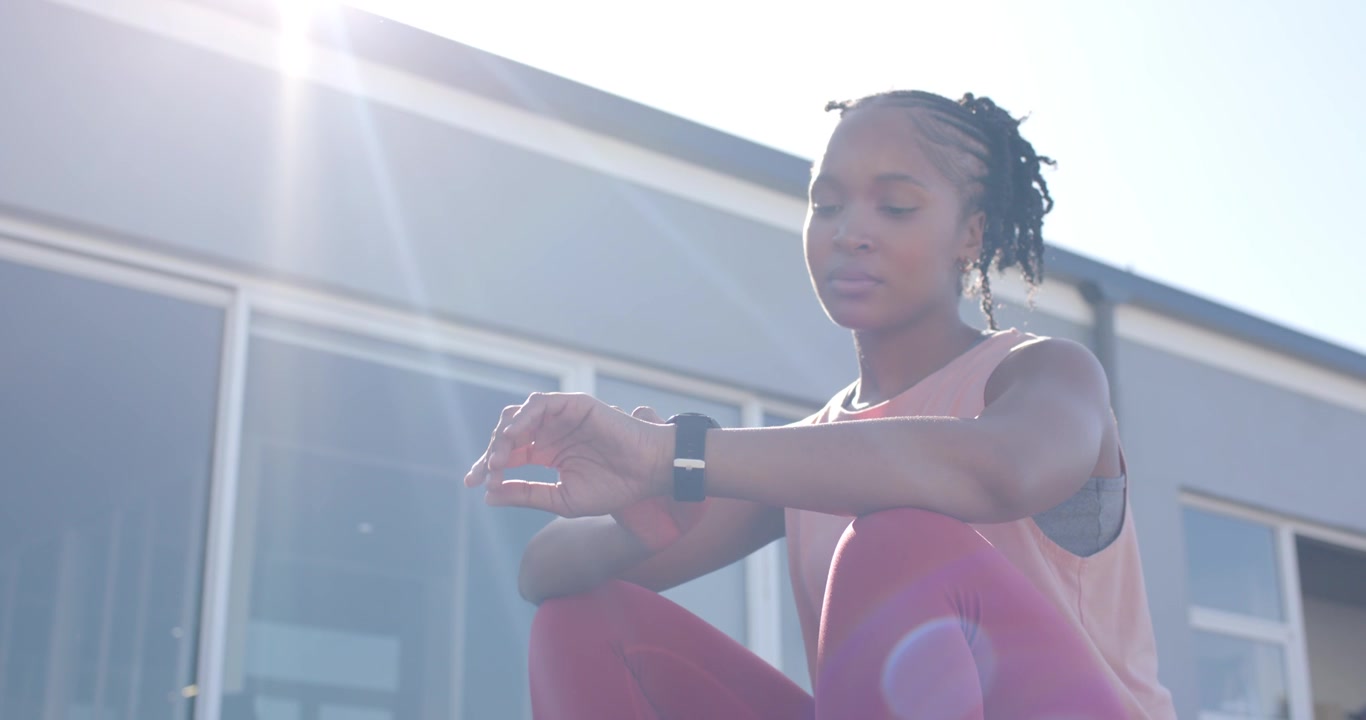 African American woman sitting on step by glass door in gym gear, sunlight causing tracker checking