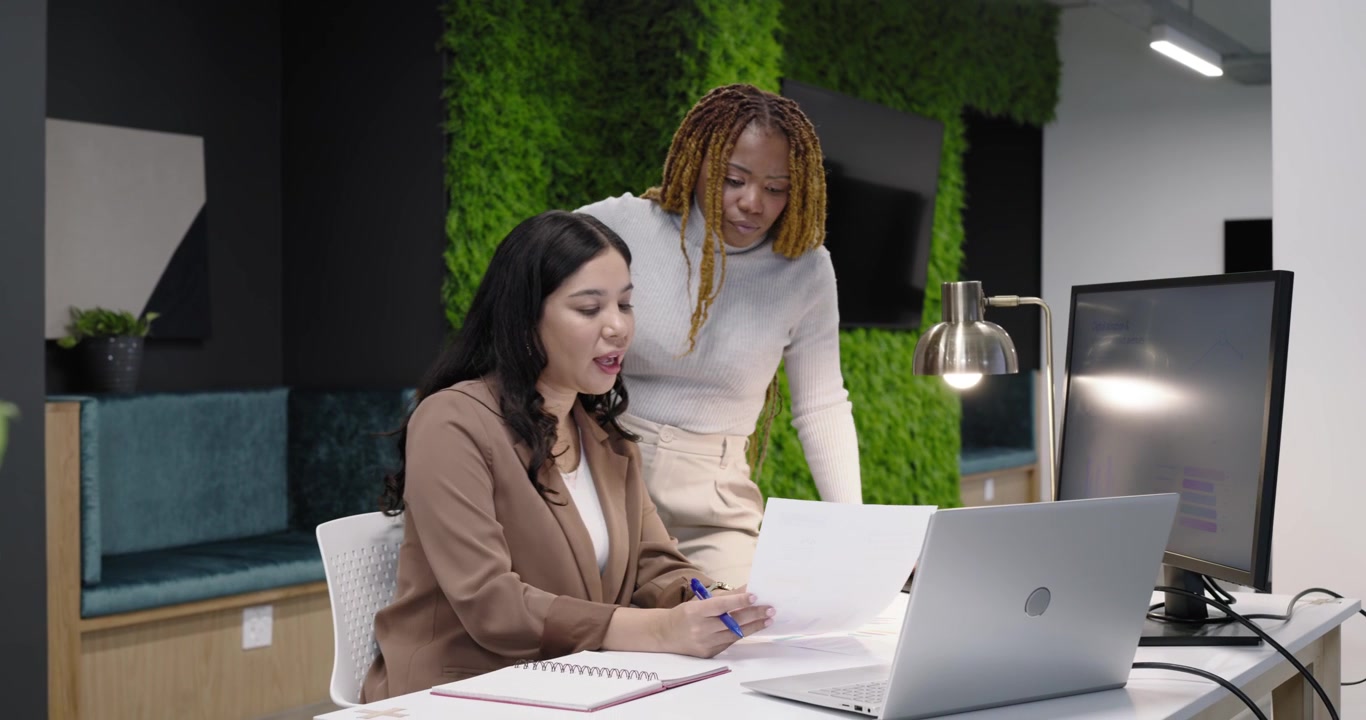Diverse female coworkers reviewing papers with blue pen as colleague approaching pointing at laptop