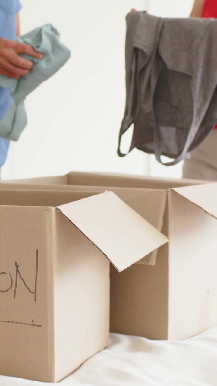 Vertical video: Holding folded shirt, man in blue passing gray tank-top into moving boxes on bed