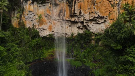 slow reveal of a towering waterfall cascading down a colourful rock cliff into a natural swimming hole