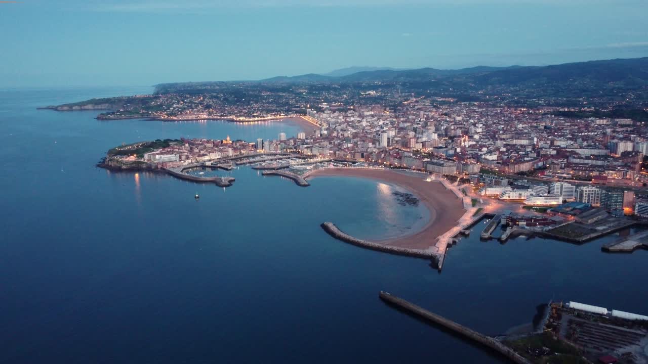 Vista aerea con drone del paesaggio urbano di Gijón illuminato al crepuscolo con traffico nel centro città e porto commerciale.