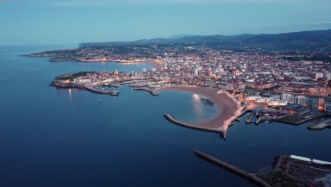 Luftdrohnenansicht-Des-Stadtbildes-Von-Gijon,-Das-In-Der-Dämmerung-Mit-Dem-Verkehr-Im-Stadtzentrum-Und-Dem-Handelshafen-Beleuchtet-Wird