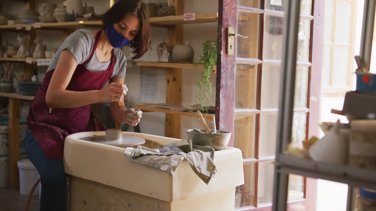 alfarera femenina con máscara facial y delantal creando cerámica en la rueda del alfarero en el estudio de cerámica