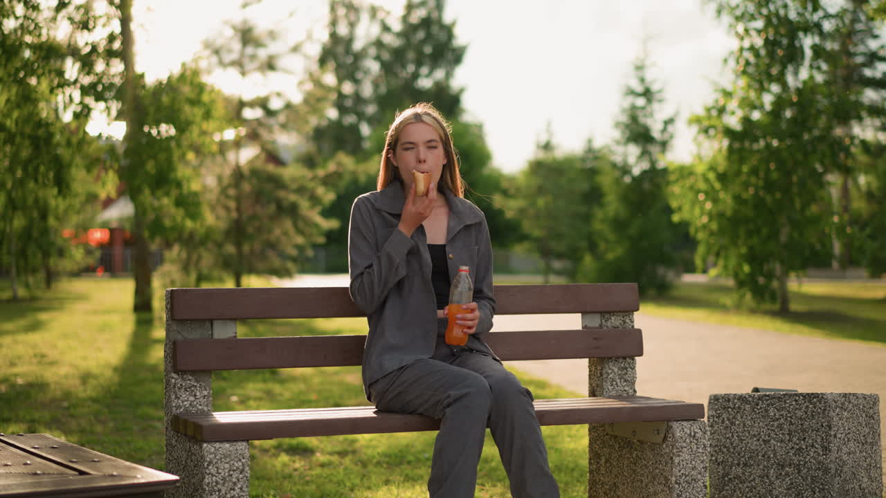 회색 옷을 입은 여자가 야외의 공원 벤치에 앉아서 간식을 먹고 술 한 병을 들고 술을 마시며, 초록색과 나무로 둘러싸여 있습니다.