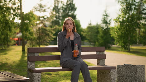 lady in grey clothing sitting on a park bench outdoors, eating a snack and holding a bottle of juice as she drinks from it, surrounded by greenery and trees in the background