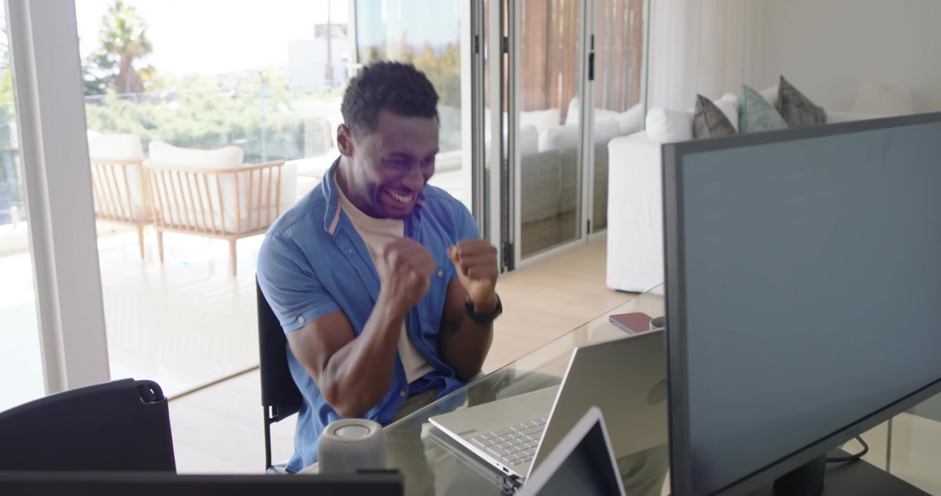 Uomo africano di mezza età alla scrivania che sorseggia da una tazza, guarda il laptop e festeggia dopo aver visto il risultato.