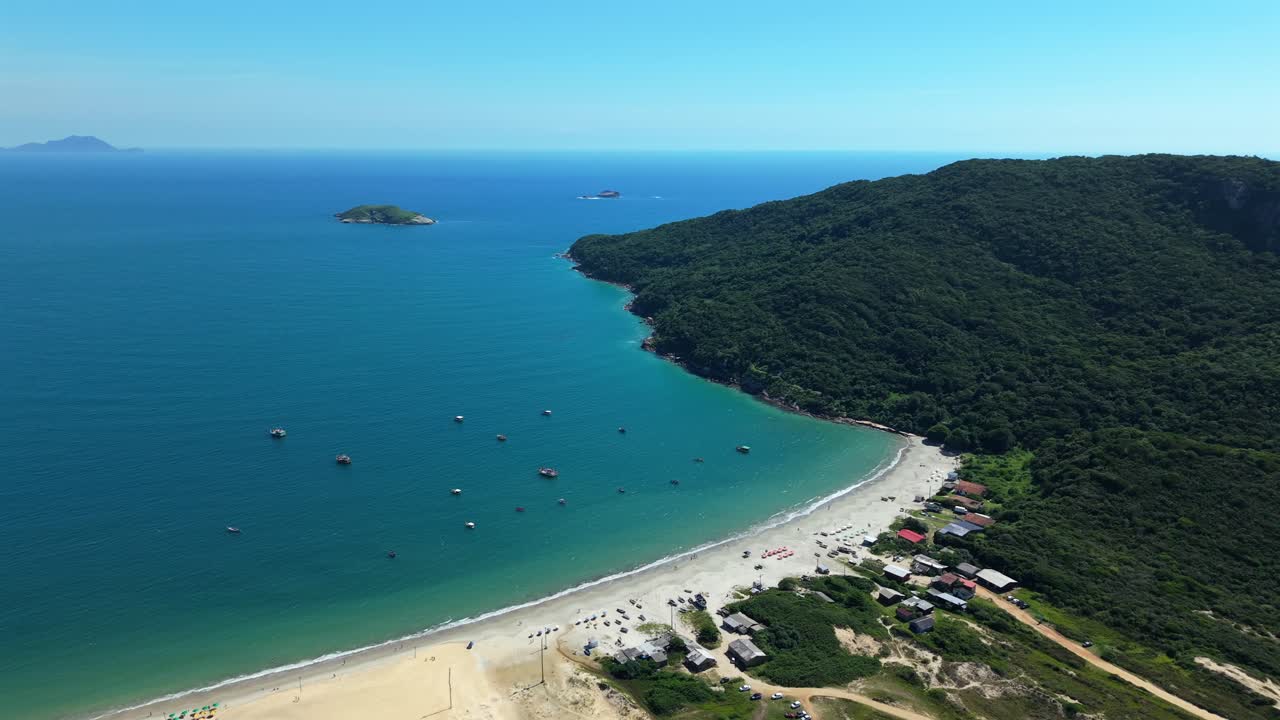 Praia dos Ingleses sulla costa nord dell'isola di Santa Catarina, Florianópolis, con una baia curva dalle acque atlantiche turchesi, un promontorio boscoso, una spiaggia sabbiosa e barche e strutture balneari sparse.