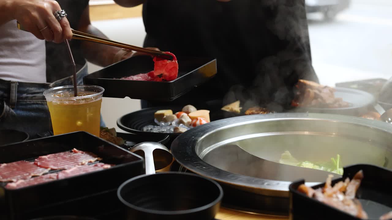 Two people add raw beef slices to steaming hotpot, indoor dining, natural daylight, close-up view