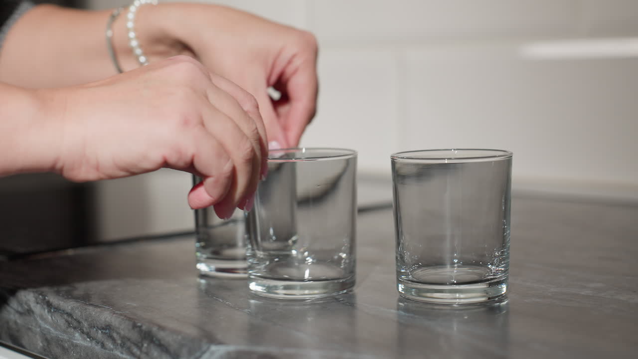 close up hand view of woman with bracelet playfully rearranging three glass cups on marble countertop, changing positions in casual yet focused movement with elegant details and smooth motion