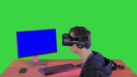 joven jugador en auriculares de realidad virtual sentado en la mesa en una pantalla verde, clave de croma