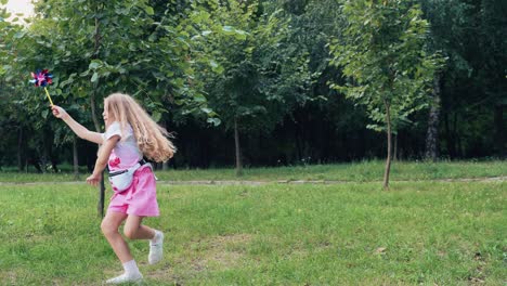 little girl with long blond hair spins, jumps and plays with a toy windmill. summer park with green grass and trees