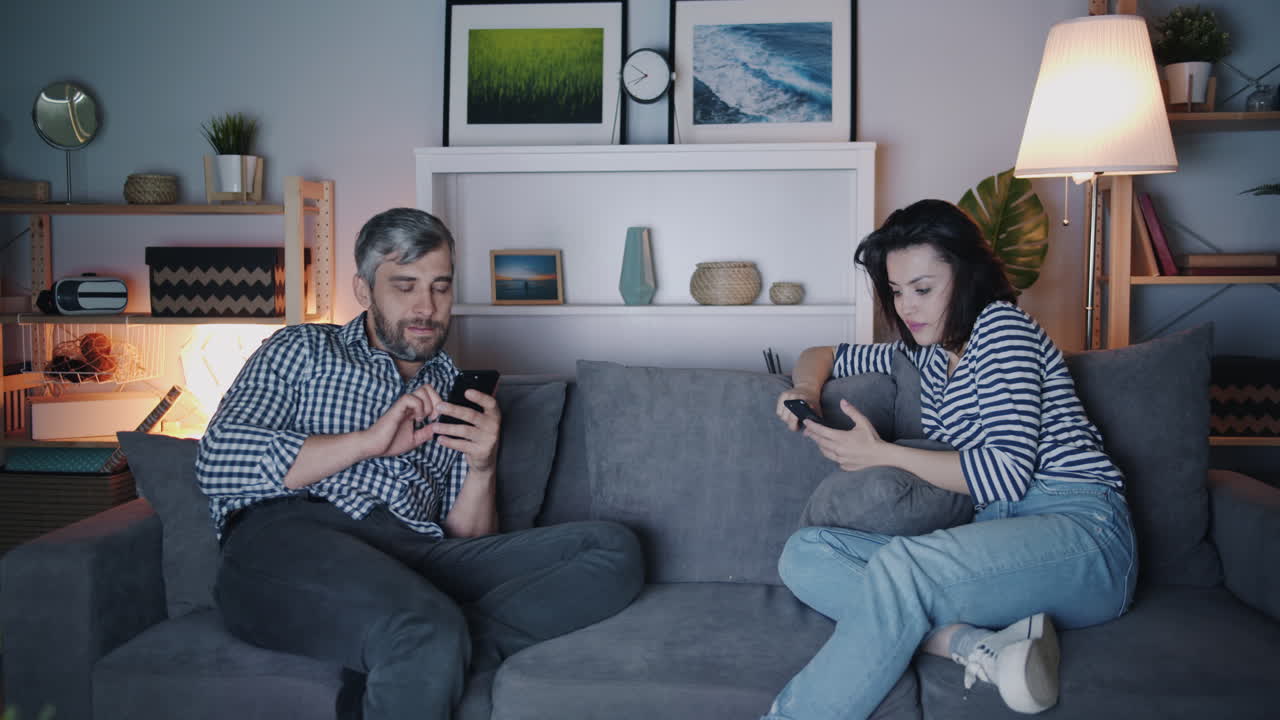 Couple relaxing on a couch at night, using smartphones