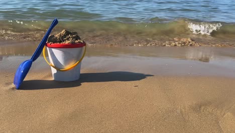 cubo de juguete lleno con cuchara azul en la playa de arena con olas rompiendo en la orilla