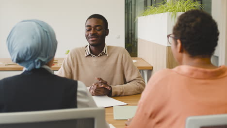 an woman and a muslim woman co workers interview a young man sitting at a table in the office 8