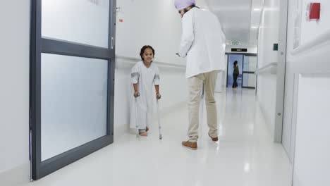 happy biracial doctor walking and talking to sick girl patient with crutches in slow motion