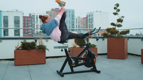 athletic girl performing aerobic training twine exercises on cycle stationary bike on house roof