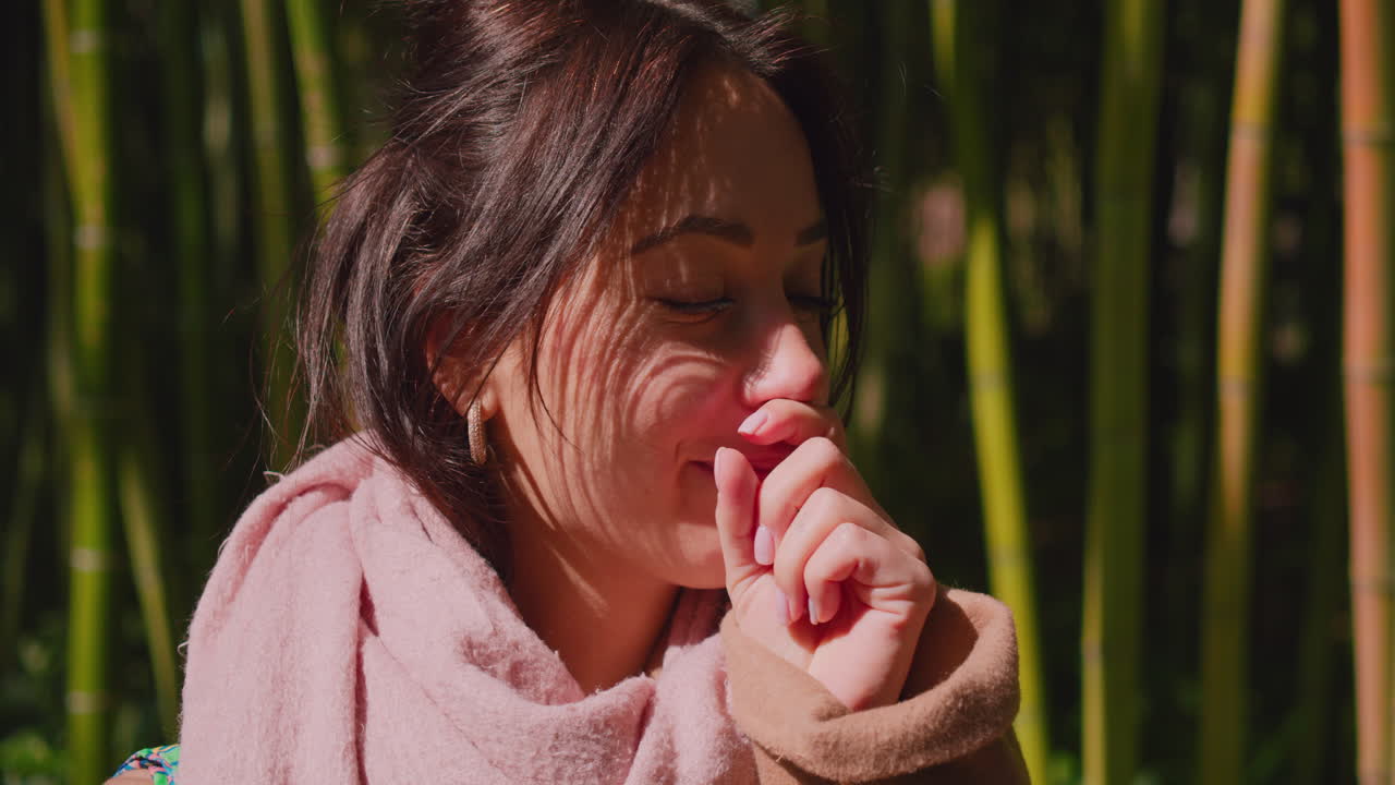 vista lateral de una mujer joven que usa una bufanda rosa de algodón suave con una tos con picazón en la garganta y se cubre la boca con una mano contra un fondo de bambú borroso