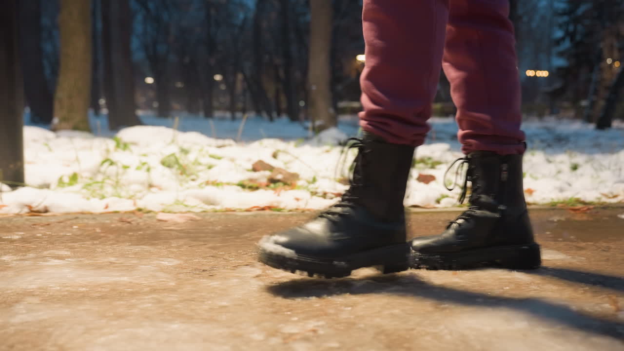 primer plano de un pie con botas pisando un camino cubierto de nieve en un parque de invierno, luces de la calle iluminadas en el fondo, mostrando nieve, clima frío y entorno invernal urbano, con suelo nevado