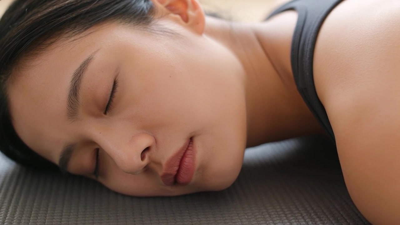 Donna asiatica sdraiata che si rilassa su un tappetino da yoga in studio mentre la telecamera registra, indossando una canotta sportiva scura.