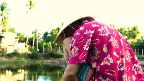 Hombre-Con-Camisa-Exótica-Y-Sombrero-Cónico-Preparándose-Para-Pescar-En-Un-Estanque-De-Agua-Local,-Soleada-Noche-Tropical